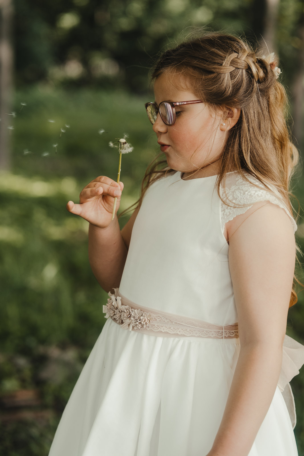 Meisje met bril in een witte communiejurk blaast een paardenbloem uit in het bos