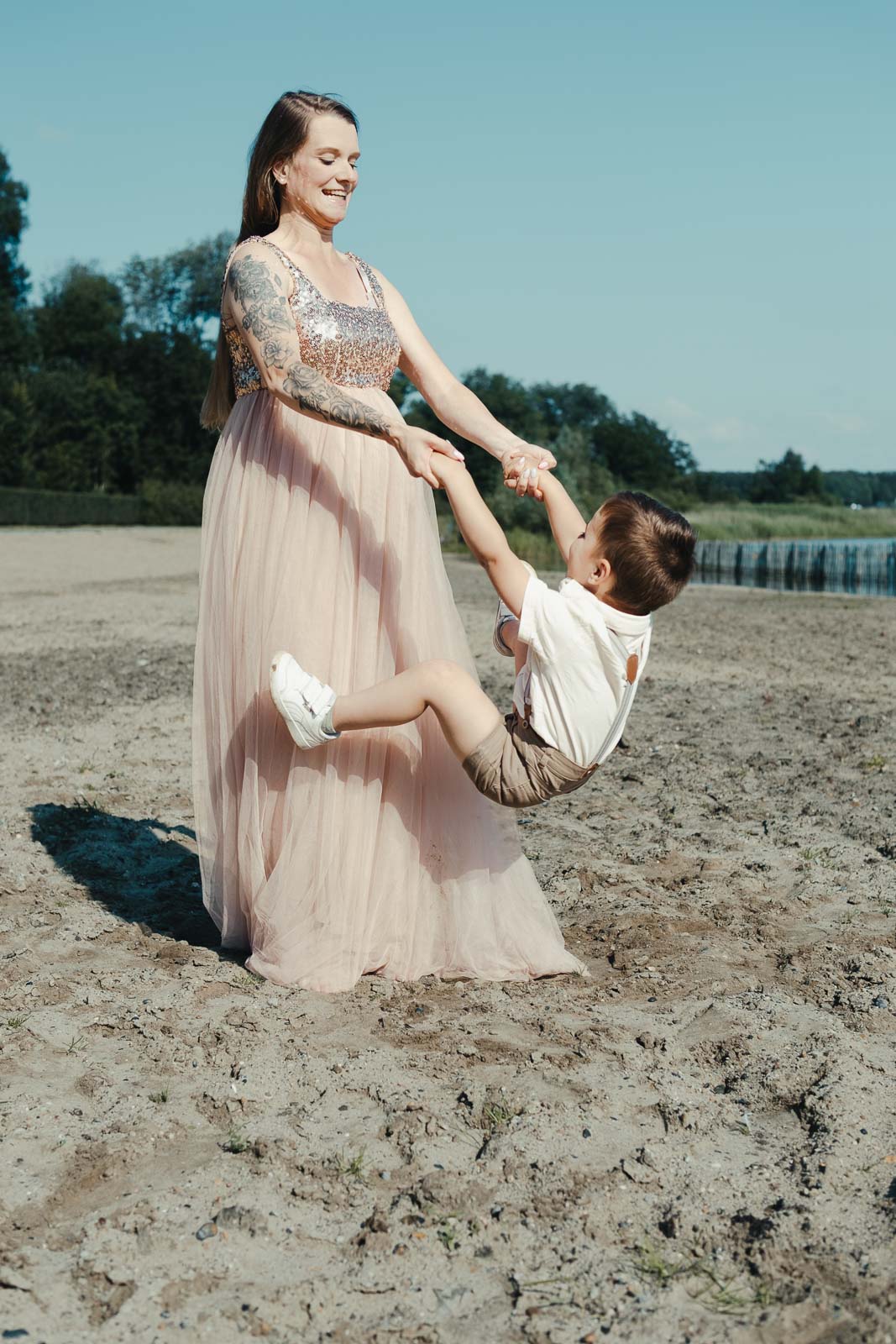 Zwangere mama in een lange roze jurk zwaait haar zoontje rond op het strand